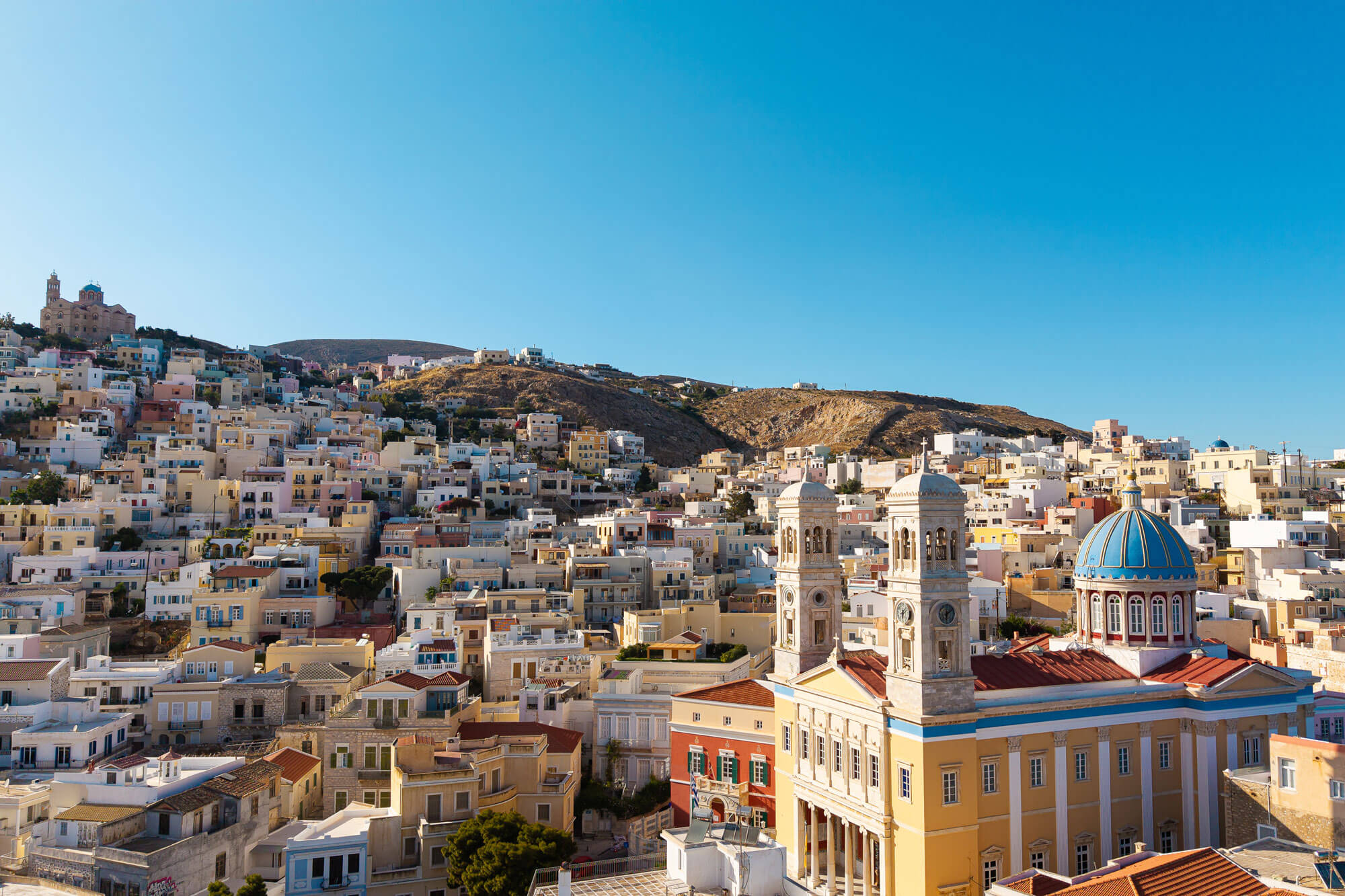 A hillside town with densely packed beige and pastel buildings, featuring a large neoclassical church with twin towers and a blue dome under a clear blue sky.