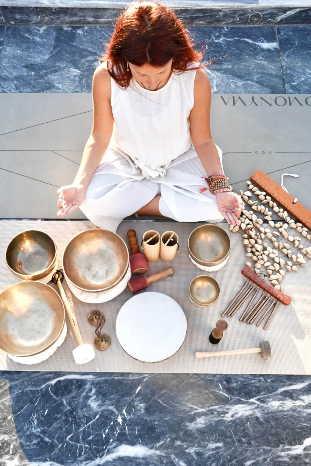 Woman with red hair in white clothes sitting cross-legged on a mat surrounded by various meditation and sound healing instruments.