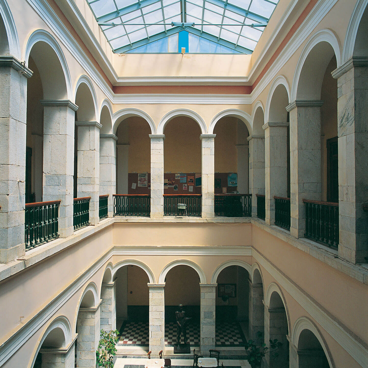 Interior courtyard of a two-story building with arched stone columns, a glass ceiling, and a statue on a black-and-white checkered floor.